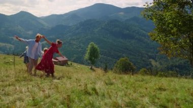Cheerful family playing active games on green grass mountain slope. Playful people running with wide open hands on meadow sunny day. Playful parents feeling happy with children summer holiday.