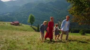 Carefree family walk together on green slope sunny day. Cheerful young couple with children feeling happy spending summer holiday in mountains. Smiling parents enjoying beautiful nature view with kids