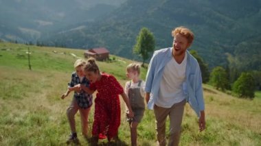 Playful parents have fun with children on grassy hill sunny day. Happy family fooling walking in beautiful mountains. Joyful couple with kids coming on top green slope summer weekend. Leisure concept.