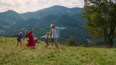 Playful family playing running like planes on green meadow. Cheerful parents with children have fun outdoor at summer holiday. Happy father mother with kids enjoying active lifestyle in mountains.