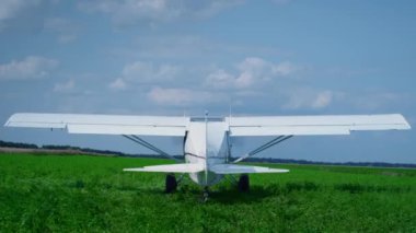 Small white airplane starting engine standing on countryside green field sunny day. Ultralight modern plane with spinning propeller preparing to flight on airfarm back view. Air transportation concept