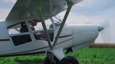 Small airplane pilot turning off engine after landing on green field. Aviation expert sitting cockpit wearing headset parking white aircraft with spinning propeller after flight in countryside.