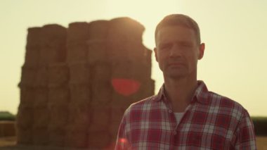 Farmer posing hay bales in sunlight portrait. Focused agronomist inspecting harvesting field. Thoughtful middle aged man agribusiness owner observing crop farmland. Agriculture professional concept.