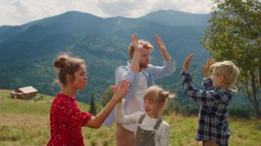 Cheerful parents giving five to joyful children on green meadow sunny day. Playful family gesticulating playing outdoors. Happy people with kids enjoying leisure in mountains summer vacation.