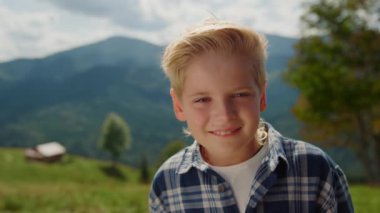 Portrait of happy boy looking camera on mountains sunny day. Carefree little man posing standing on green meadow close up. Positive cute preteen guy spending summer holiday on nature. Leisure concept.