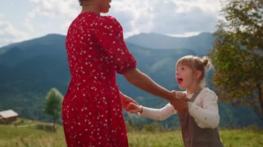 Side view of cheerful mother jumping with daughter dancing on mountain hill close up. Smiling woman with cute girl holding hands bouncing outdoor. Happy mom having fun with child at summer holiday.