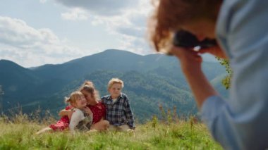 Back view of red hair father taking photos mother with children sitting green grass mountain meadow. Smiling family posing camera on nature sunny day. Happy people enjoy summer holiday together.