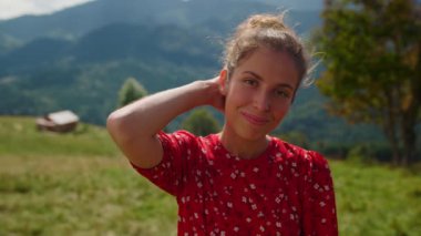 Closeup young attractive woman looking camera on green meadow. Portrait of positive girl smiling outdoors enjoying sunny day. Happy lady wearing red dress touching hairstyle standing mountains slope.