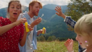 Cheerful family enjoy blowing soap bubbles outdoor for catching children close up. Smiling mother father have fun summer holiday in mountains. Happy couple playing with carefree kids on green meadow.