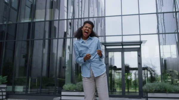 Joyful happy woman celebrating victory at modern office center. Triumph moment. Cheerful attractive African American girl jumping raising hands up as get job promotion. Energetic winner shouting loud.