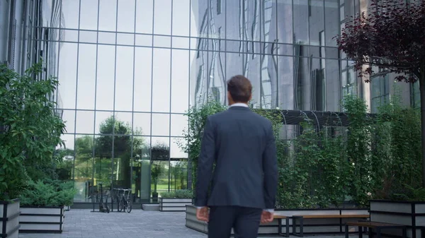 Confident businessman walking downtown area rear view. Modern glass office building facade with neat landscaped garden. Successful man director going workplace in suit. European financial institution.