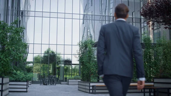 Confident businessman walking business center garden alone. Success concept. Unknown man financial director going to workplace empty landscaped area in suit. Relaxed manager stroll downtown rear view