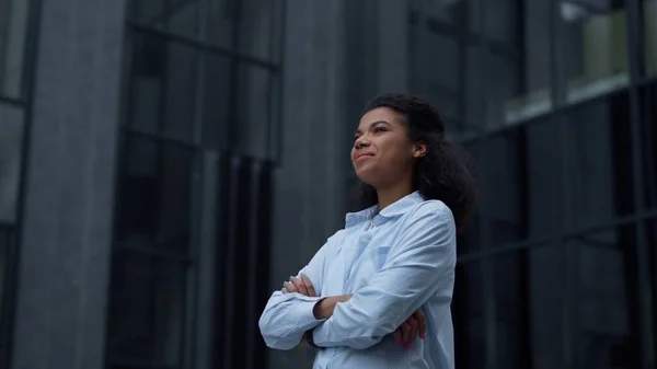 Proud woman standing alone outdoors portrait. Girl pose inspect modern building. Cheerful winner with crossed hands looking in camera. Attractive african american manager enjoying moment of triumph.