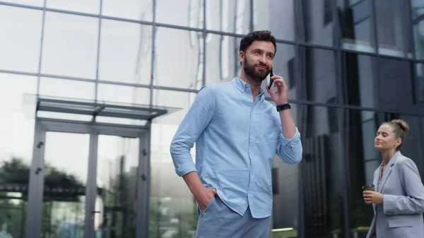 Handsome man speaking cellphone at glass office building. Modern corporate area. Diverse employees passing by at downtown. Joyful bearded entrepreneur having phone conversation with business partner