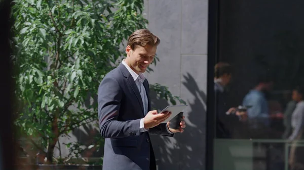 Smiling finance worker checking smartphone walking in modern workplace outside. Cheerful business man stepping holding coffee cup in contemporary office yard portrait. Relaxing lunch break concept.