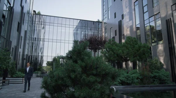 Confident businessman walking office building. Landscaped garden at downtown. Handsome man successful manage go peaceful green recreation spot at business center. Modern finance corporation exterior