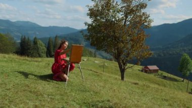 Young inspired girl painter creating picture on green hill sunny day. Talented woman painting artwork on easel using paintbrush outdoors. Creative artist making sketch of summer mountains at holiday.