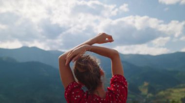 Unknown girl standing raising hands over head in mountains summer time close up. Back view of relaxed woman enjoying cloudy landscape outdoors. Young lady wearing red dress spending holiday on nature.