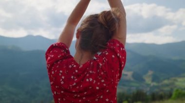 Unrecognizable woman relaxing raising hands to cloudy sky outdoors close up. Back view of young girl enjoying summer vacation in mountains. Unknown lady wearing red dress rest on nature alone.