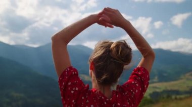 Young woman admiring mountains landscape enjoying summer holiday close up. Back view unknown girl raising hands standing alone on green hill. Unrecognizable lady wearing red dress enjoying nature.