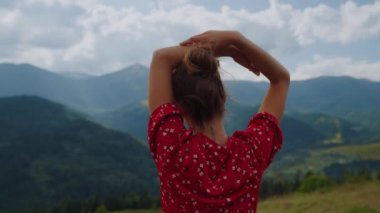 Back view of unknown tranquil woman standing in front mountains scenery close up. Relaxed girl raising hands over head wearing red dress. Pretty lady enjoying summer holiday on cloudy nature.