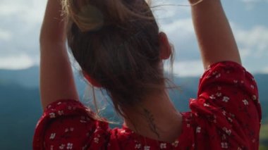 Back view of raising hands young woman resting in summer mountains close up. Gentle girl arms touching each other in front cloudy sky. Unrecognizable model enjoying nature standing beautiful meadow.