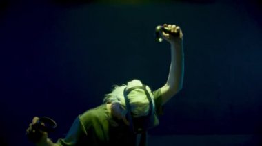 Man playing vr headset game in neon lights. Excited gamer gesturing holding controllers. Unrecognized blonde guy selecting virtual objects in simulation. Energetic metaverse teenager experiencing ar.