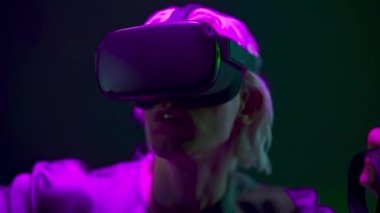 Amazed man playing videogame with remote controllers closeup. Focused gamer using joysticks and VR goggles at neon light place. Blonde headset guy interacting digital interface alone. Future concept 