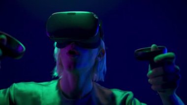 Energetic gamer playing online videogame with gamepads closeup. Active vr glasses man enjoying virtual reality with controllers. Blonde guy using joysticks for quest adventure game at neon light