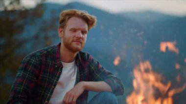 Adventurous man look fire burning on cozy evening. Close up young camper rest in mountains landscape. Positive guy enjoy relax on hiking trip. Smiling traveler rest on camp journey. Outdoors concept.