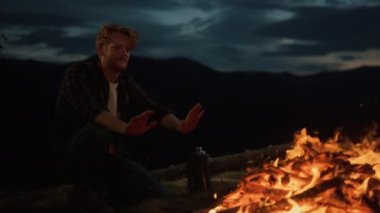 Dreamy hipster enjoy night fire burn on camp site. Closeup young traveler relax in mountains nature. Lonely millennial warm hands by campfire in dusk. Chill tourist rest on journey. Leisure concept.
