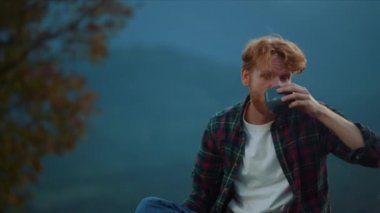 Tired camper relax drink tea in mountains nature sunset. Handsome hipster enjoy campfire outside. Hiker guy spend holiday on journey close up. Thoughtful traveler sit alone on trip. Travel concept.