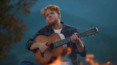Closeup musician enjoy evening in mountains. Hipster guy play guitar on nature burning campfire. Happy male hiker rest by bonfire outdoors. Red hair millennial spend vacation on camp. Youth concept.