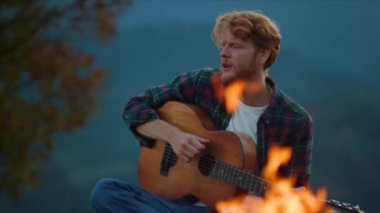 Young guitarist play outdoors on evening camp. Closeup carefree guy enjoy guitar music in forest. Redhead tourist sing song by burning campfire on nature. Smiling hiker rest on travel. Leisure concept