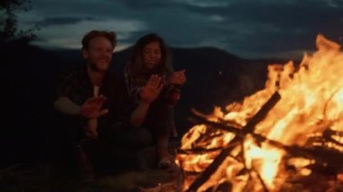 Close up two travelers enjoy bonfire on evening nature. Resting couple warm hands on campfire outdoors. Smiling lovers spend vacation time together on dark night dusk. Relax tourism journey concept.