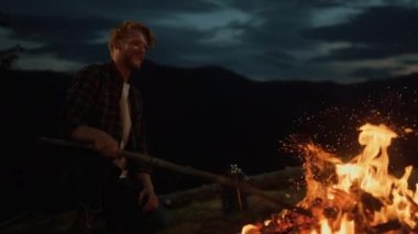Joyful man camping outside on dark evening. Close up mountains traveler relax on nature alone. Adventurous millennial enjoy watch fire burning on campfire. Recreation freedom wanderlust youth concept.