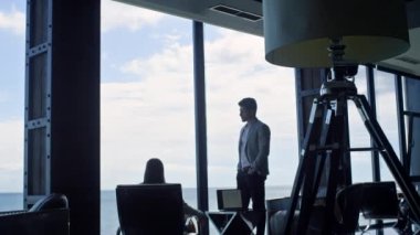 Business couple silhouettes thinking at marine view. Two unknown coworkers discussing business deal at panorama window. Unrecognized partners having conversation in dark interior conference room 