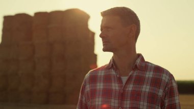Farm worker golden sunlight at hay bales closeup. Pensive man observing harvest field on sunset. Successful agronomist inspecting wheat farmland on autumn day. Agribusiness owner professional concept.