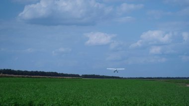 Small ultralight airplane takeoff from green grass farmland. White propeller plane accelerating speed flying on airfield against blue cloudy sky. Professional aviator riding modern air transport.