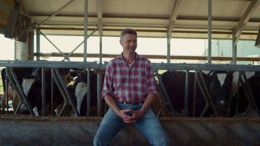 Farm worker relax after hard day sitting barn. Agronomist man enjoying work feeding cows in modern cowshed. Happy agricultural specialist posing on camera near cattle animals. Farming rest concept.