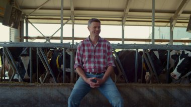 Happy farmer posing sitting on cowshed feeder near healthy cows. Charming professional agro worker enjoying work process in farmland barn. Middle age ranch owner smiling relaxing on modern farm.