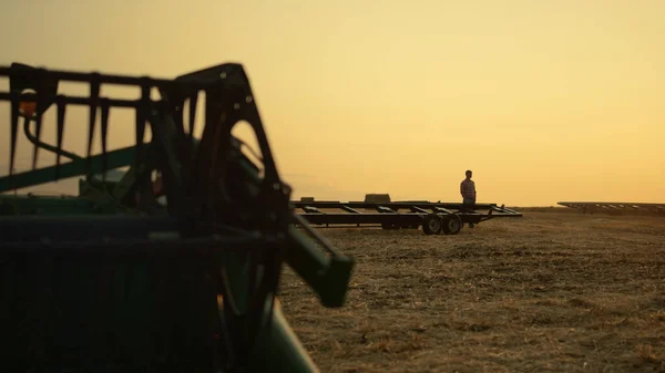 黄金の日没時にコムギ畑の収穫装置で農家のシルエット シリアル農地に立っている未知の農業経営者は クロップ機械の作業日の終わりを検査する アグロコンセプト — ストック写真