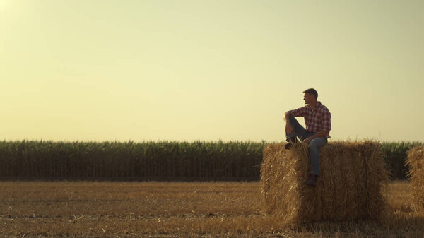 Farmer resting hay stack in autumn evening. Focused worker inspecting harvest field in autumn. Thoughtful man businessman consider crop planning relax on wheat straw bale. Rural lifestyle concept.