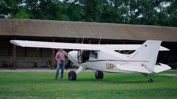 Aviator inspecting small white airplane landed on countryside airdrome. Professional man aviator checking private plane parked on green grass airfield with open door waiting flight. Aviation concept