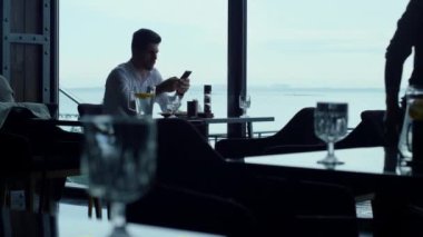 Successful entrepreneur working on smartphone at breakfast time in stylish sky bar. Busy employee sending emails at lunch break in restaurant. Unrecognizable male silhouette walking hotel cafe hall.