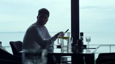 Handsome man scrolling smartphone touchscreen in modern restaurant. Thoughtful silhouette freelancer using cellphone device indoors. Elegant businessman resting luxury cafe. Busy lifestyle concept.