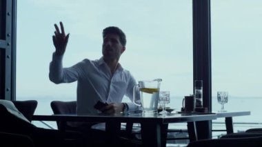 Successful businessman customer asking dinner bill gesture in luxury cafe. Confident ceo manager calling waiter while resting panoramic bar terrace. Focused man talking mobile phone in restaurant.