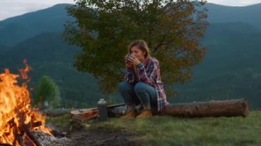 Lonely traveler rest forest in mountains. Relaxed woman drink tea by campfire outdoors close up. Thinking girl enjoy holiday vacation on nature tourism activity. Wanderlust journey camp trip concept.