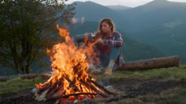 Camp woman relax mountains. Close up beautiful girl sit log enjoy bonfire on nature vacation. Young traveler drink thermos tea on picnic outdoors. Thoughtful tourist rest in forest. Camping concept.