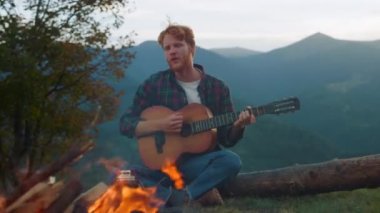 Signing man relax nature on tourism holiday. Closeup ginger millennial enjoy campfire on mountains. Carefree guy play acoustic guitar on evening nature. Happy backpacker enjoy outside. Travel concept.